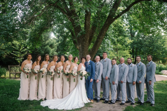 A wedding party poses outdoors under a large tree. The bride in a white gown and groom in a blue suit stand center, surrounded by bridesmaids in pale dresses and groomsmen in light gray suits, all holding bouquets and smiling. by Justin Salem Meyer