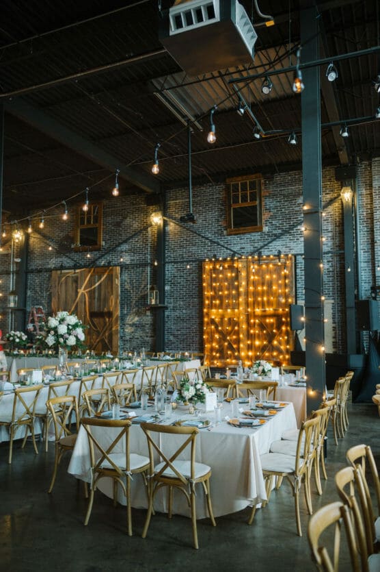 A stylish indoor wedding reception features long tables with white linens, wooden chairs, floral centerpieces, and string lights draped across brick walls and large barn doors, creating a warm, festive atmosphere. by Justin Salem Meyer
