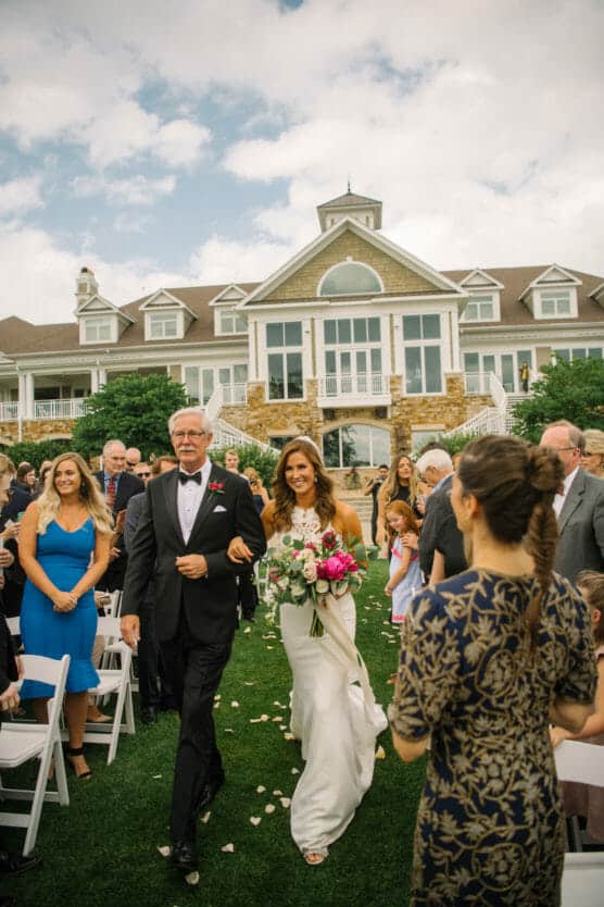 A bride in a white dress walks down an outdoor aisle with an older man, surrounded by seated guests, toward a large house. She holds a colorful bouquet and smiles as people watch. Petals are scattered on the grass. by Justin Salem Meyer