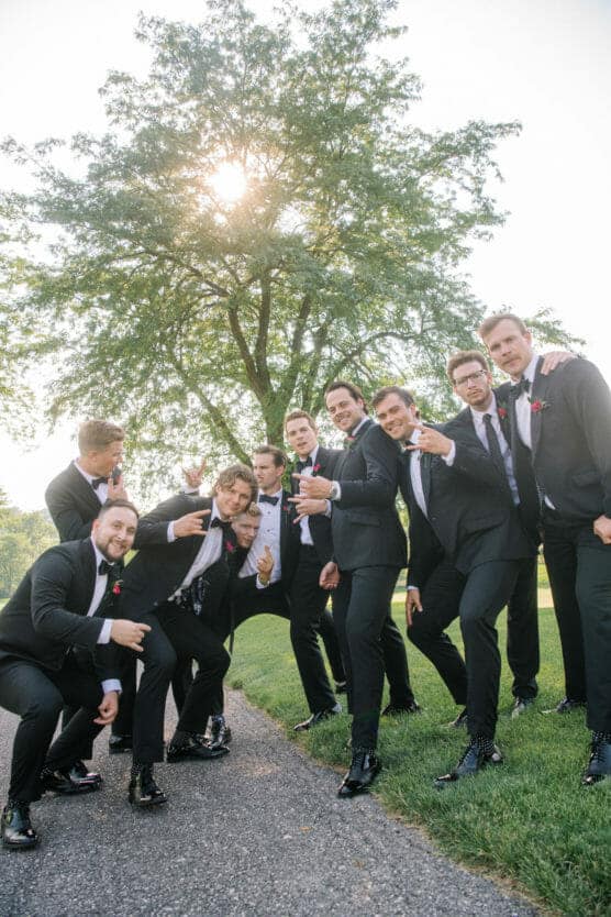 A group of men in black suits and bow ties pose playfully outdoors on a sunny day, with a large tree and sunlight in the background. They smile and gesture towards the camera on a grassy area by a path. by Justin Salem Meyer