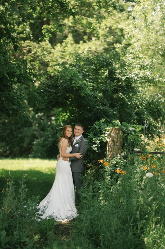 A bride in a white dress and a groom in a suit stand together, smiling, surrounded by lush green trees and wildflowers in an outdoor garden setting. by Justin Salem Meyer