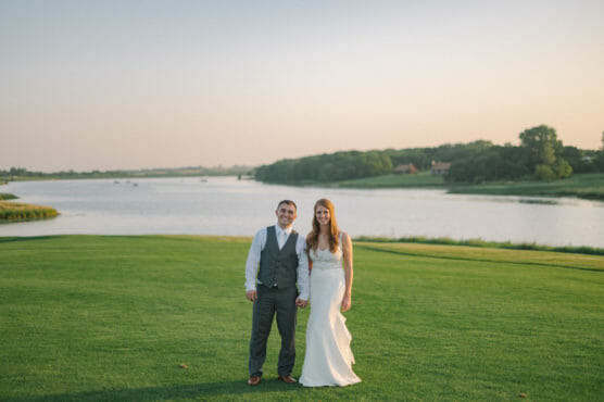 A bride and groom stand hand in hand on a green lawn by a lake at sunset, both smiling with trees and calm water in the background. by Justin Salem Meyer