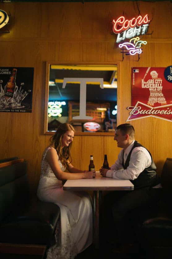 A bride and groom sit in a booth at a bar, smiling and holding beer bottles. Neon signs and posters decorate the wood-paneled walls behind them, creating a casual, relaxed atmosphere. by Justin Salem Meyer