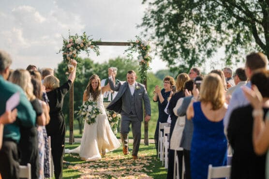A bride and groom walk hand in hand down the aisle outdoors, smiling and raising their arms in celebration as wedding guests on both sides applaud and cheer. Flower petals are scattered on the grass and a floral arch is behind them. by Justin Salem Meyer