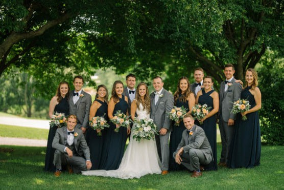 A wedding party poses outdoors under green trees. The bride in a white dress and the groom in a gray suit stand in the center, surrounded by bridesmaids in dark dresses and groomsmen in gray suits, all holding bouquets and smiling. by Justin Salem Meyer
