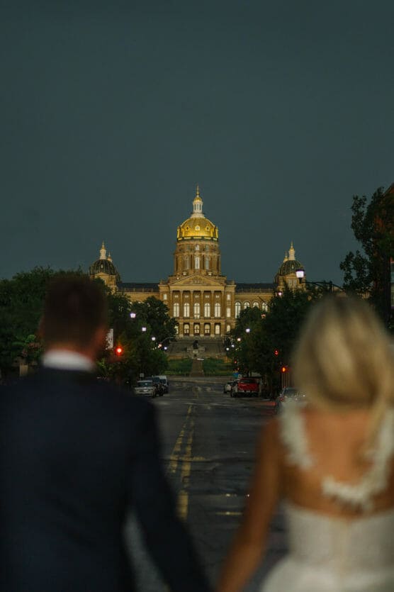 A couple, seen from behind and holding hands, faces a grand, illuminated capitol building at dusk on an empty, wet street. by Justin Salem Meyer
