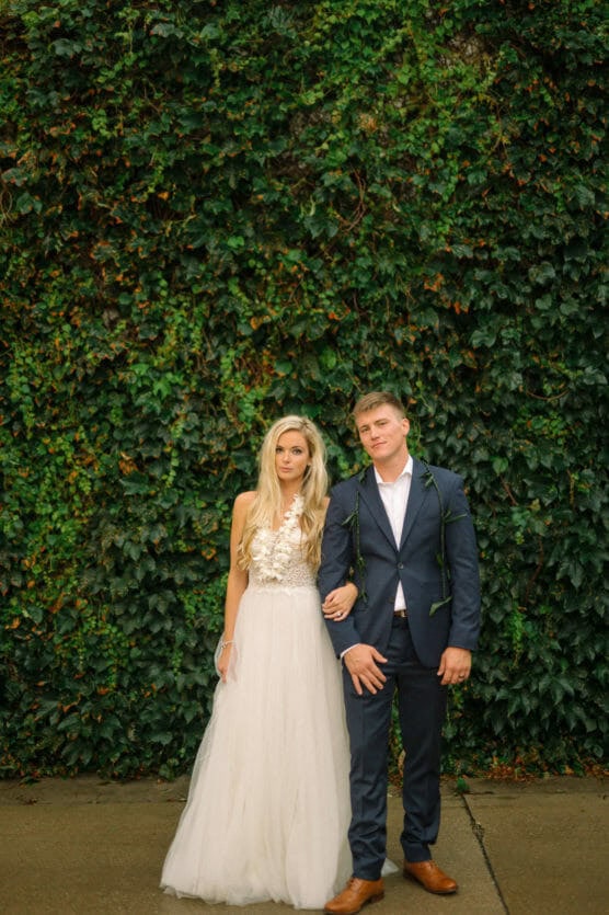 A bride in a white dress and a groom in a navy suit stand together against a lush green ivy-covered wall, holding hands and looking at the camera with neutral expressions. by Justin Salem Meyer