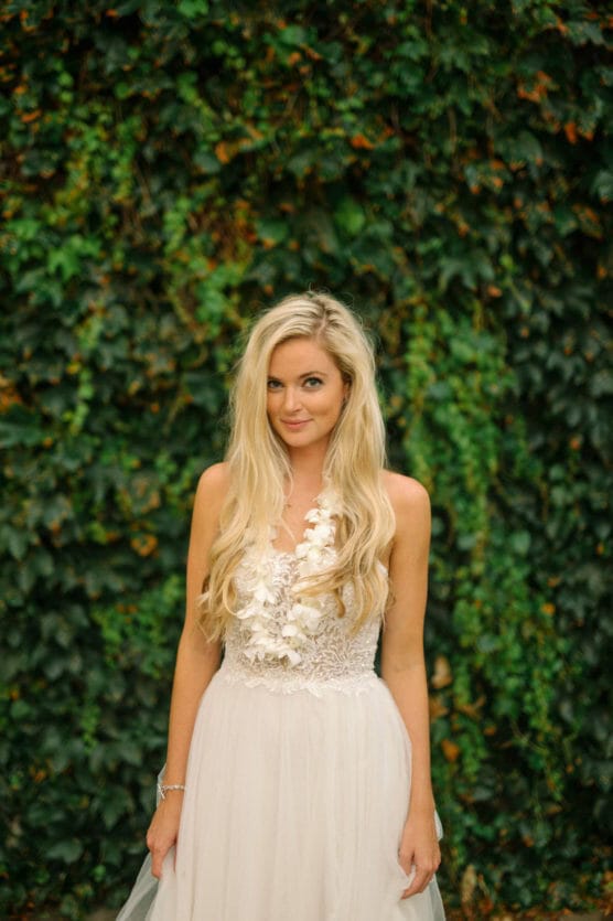 A woman with long blonde hair in a white lace dress stands in front of a lush wall covered in green ivy, looking at the camera with a gentle smile. by Justin Salem Meyer