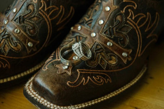 A pair of ornate brown cowboy boots with detailed stitching and silver studs, with a diamond engagement ring and a wedding band resting on the toe of one boot. by Justin Salem Meyer