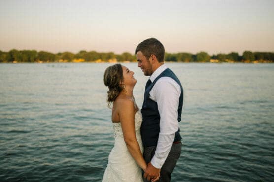 A bride and groom stand by the water, facing each other and holding hands, both smiling. The background features a calm lake and distant trees under a clear sky at sunset. by Justin Salem Meyer