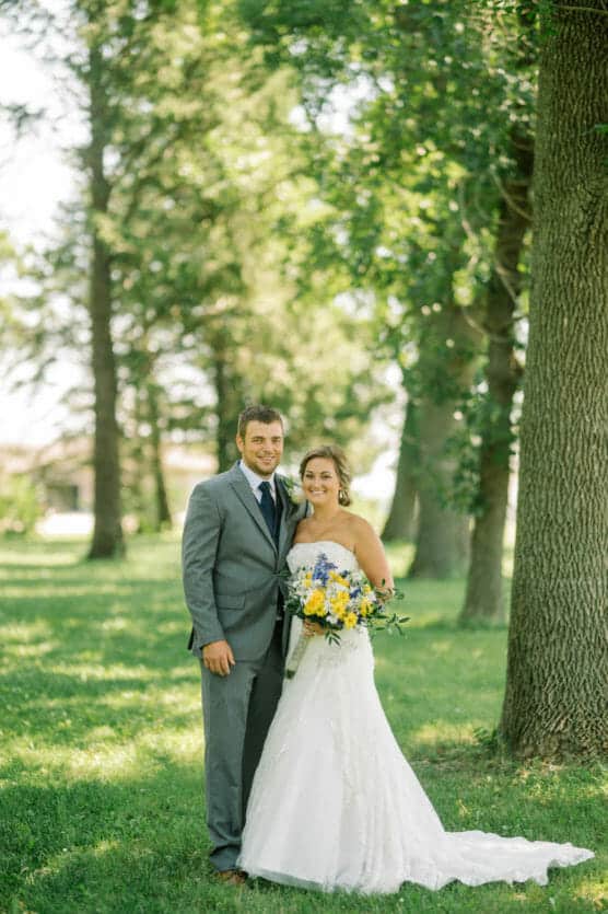 A bride in a white dress holding a bouquet of yellow and blue flowers stands beside a groom in a gray suit and tie. They are smiling outdoors in a sunlit, green park with trees in the background. by Justin Salem Meyer