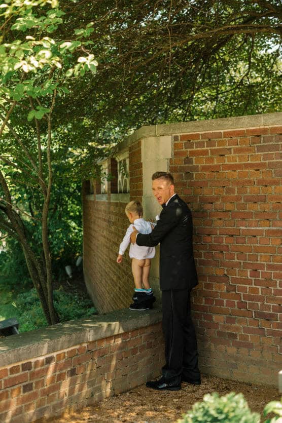 A man in a black suit helps a young child balance while standing on a brick wall, surrounded by lush greenery and sunlight filtering through tree branches. by Justin Salem Meyer