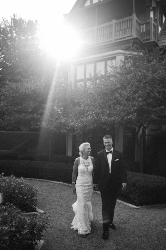 A bride and groom walk hand in hand outside, smiling at each other, with sunlight streaming down beside a large house and lush trees in the background. by Justin Salem Meyer