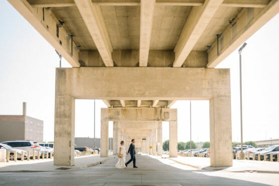 A bride and groom walk hand-in-hand under a large, concrete overpass on a sunny day, surrounded by parked cars and industrial buildings, with sunlight streaming through the structure. by Justin Salem Meyer