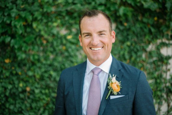A man in a blue suit with a pink tie and yellow boutonniere smiles in front of a lush green ivy-covered wall. by Justin Salem Meyer