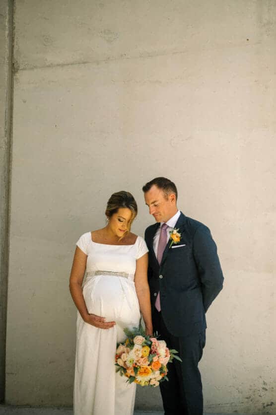 A pregnant bride in a white dress holds her belly and a bouquet of flowers while standing beside a groom in a suit with a floral boutonniere, both looking downward against a plain, light-colored wall. by Justin Salem Meyer