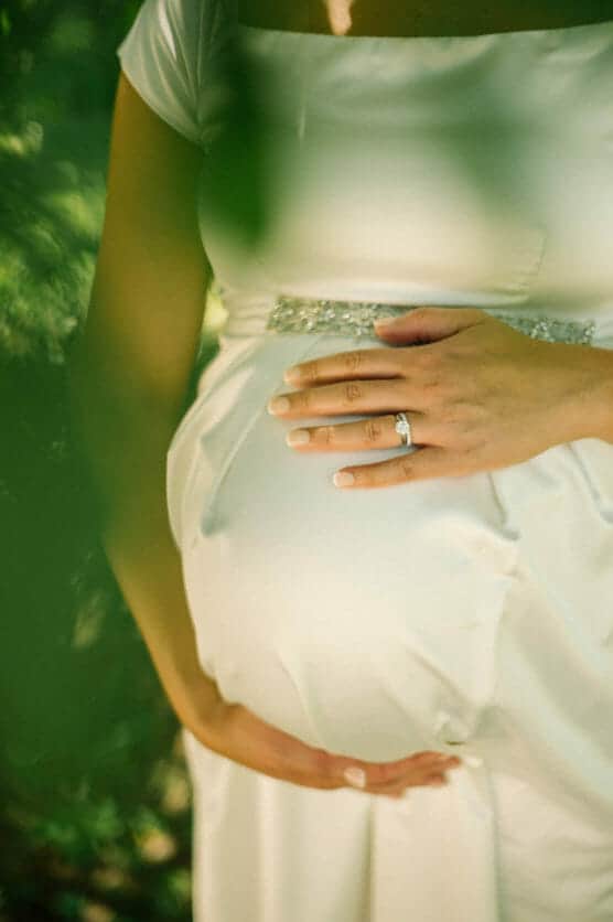 A person in a white dress cradles their pregnant belly with both hands, showing a wedding ring on one hand. The image is softly lit with green foliage partially in the foreground. by Justin Salem Meyer