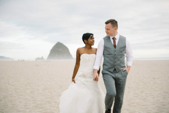 A newlywed couple walks hand in hand on a sandy beach, both smiling. The bride wears a strapless white gown, and the groom wears a gray suit vest with a tie. A large rock formation is visible in the background. by Justin Salem Meyer