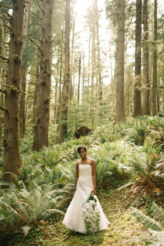 A bride in a white gown holds a bouquet and smiles while standing on a lush, green forest path surrounded by tall trees and ferns, with sunlight streaming through the branches above. by Justin Salem Meyer