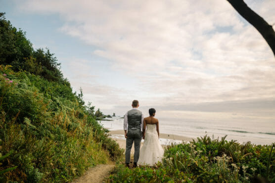 A bride and groom stand on a grassy path overlooking the ocean, holding hands and gazing at the horizon under a cloudy sky. Lush greenery surrounds them, and waves meet the sandy shore below. by Justin Salem Meyer