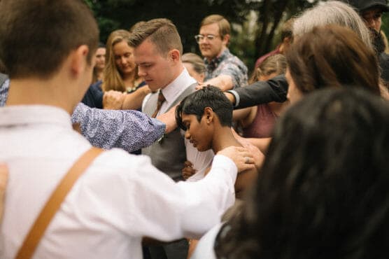 A group of people stand closely together outdoors, forming a supportive circle with their arms around each other, appearing to share a moment of comfort and solidarity. by Justin Salem Meyer