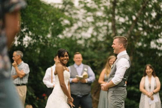 A bride and groom stand outdoors, smiling and laughing during their wedding ceremony, surrounded by wedding guests and greenery in the background. by Justin Salem Meyer