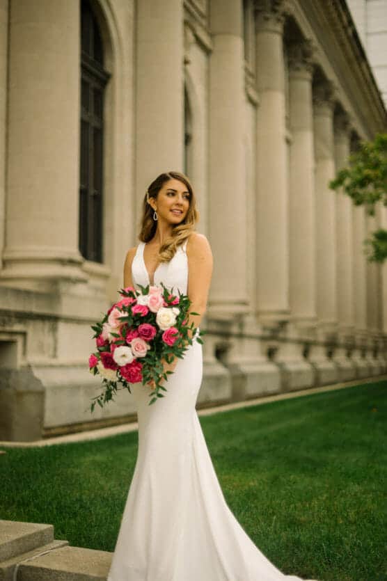 A bride in a white gown holds a large bouquet of pink and white roses, standing outside a classical stone building with tall columns and lush green grass. by Justin Salem Meyer