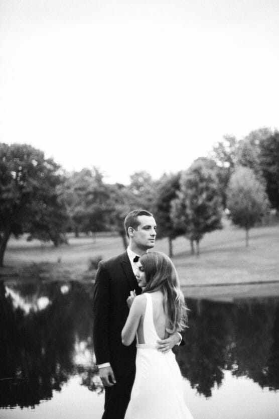 A bride in a white dress embraces a groom in a black suit by a calm lakeside, with trees and grassy fields in the background; the image is in black and white. by Justin Salem Meyer