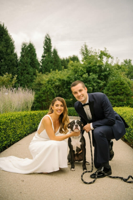 A bride in a white dress and a groom in a tuxedo crouch and smile beside a black and white dog on a path in a green garden. by Justin Salem Meyer