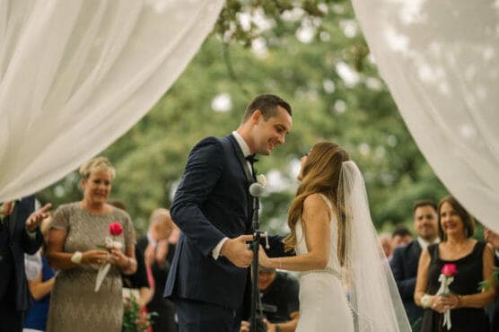 A bride and groom stand facing each other under white drapes during an outdoor wedding ceremony, surrounded by smiling guests. The groom is in a navy suit; the bride wears a veil and holds his hand. by Justin Salem Meyer