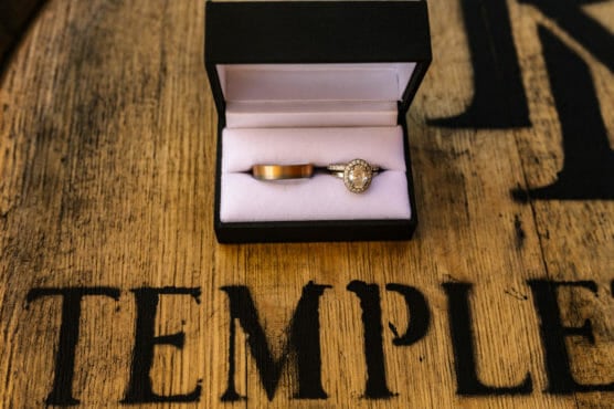 A gold wedding band and a diamond engagement ring in a black ring box, resting on a wooden surface with the word TEMPLE partially visible in bold black letters. by Justin Salem Meyer