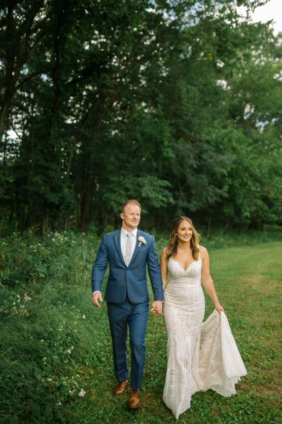 A bride in a patterned white dress and a groom in a blue suit walk hand in hand on a grassy path, surrounded by green trees, both smiling warmly. by Justin Salem Meyer