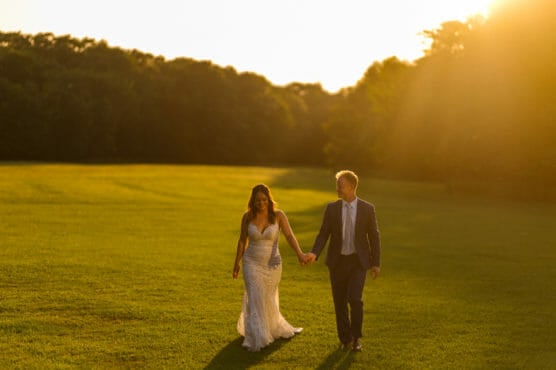 A bride in a white dress and a groom in a suit walk hand in hand across a sunlit grassy field, with trees in the background and warm golden sunlight illuminating the scene. by Justin Salem Meyer