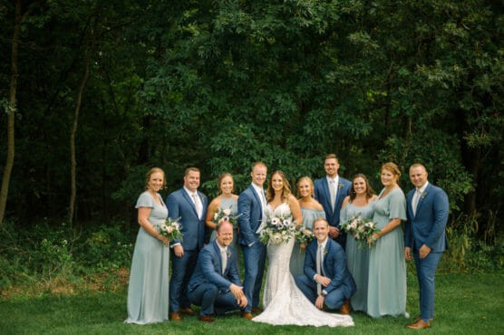 A bride and groom stand together outdoors, surrounded by bridesmaids in light blue dresses and groomsmen in blue suits, all smiling for a wedding portrait in front of a lush green forest background. by Justin Salem Meyer