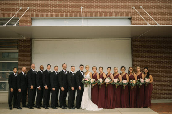 A wedding party poses in front of a brick wall. The bride stands in the center in a white dress with the groom beside her in a black suit. Bridesmaids in burgundy dresses and groomsmen in black suits stand on either side. by Justin Salem Meyer
