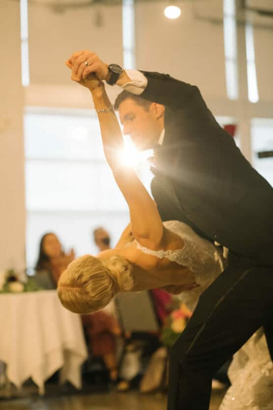 A groom in a black suit dips his bride, who wears a white lace dress, during a dance. Light shines between them, and blurred guests watch in the background. by Justin Salem Meyer
