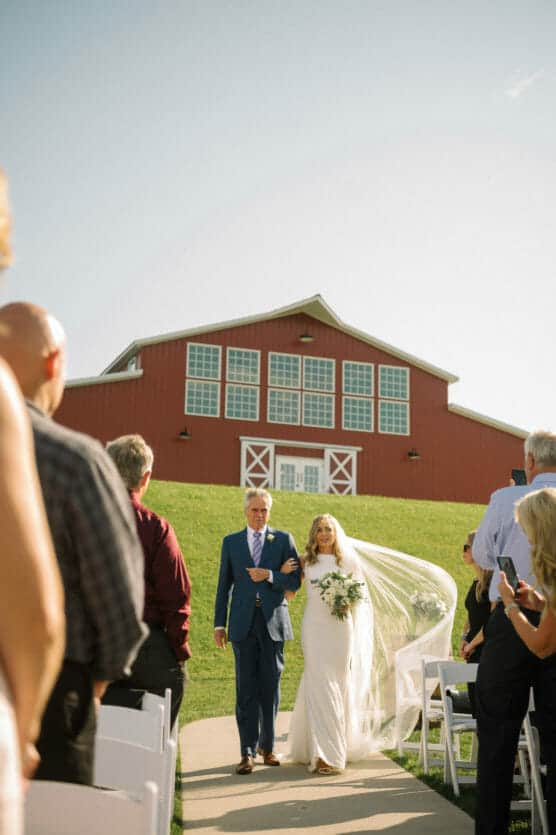 A bride in a white dress and veil walks arm-in-arm with an older man in a blue suit down an outdoor aisle, with guests seated on either side and a large red barn in the background under a clear sky. by Justin Salem Meyer