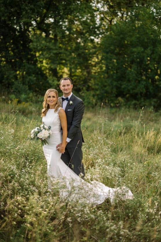 A bride in a white dress holding a bouquet stands in front of a groom in a suit, both smiling, in a grassy field with green trees in the background. by Justin Salem Meyer