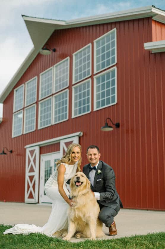 A bride and groom in wedding attire kneel and smile beside their golden retriever in front of a large red barn with white trim and tall windows on a sunny day. by Justin Salem Meyer