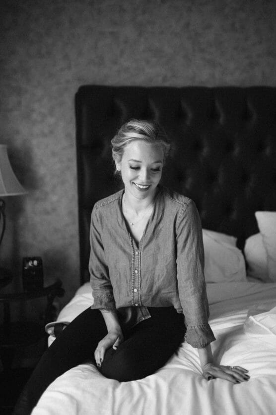 A woman with blonde hair, tied back, sits on a bed smiling and looking down. She is wearing a button-up shirt and dark pants. The room has a tufted headboard, a lamp, and pillows on the bed. The photo is black and white. by Justin Salem Meyer