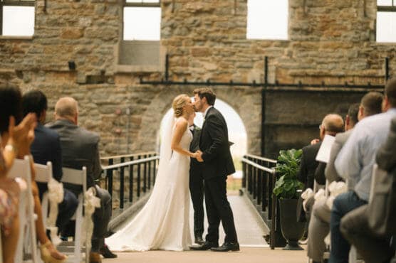 A bride and groom share a kiss at the altar during an indoor wedding ceremony, surrounded by seated guests in rows. The venue features stone walls and large windows. by Justin Salem Meyer
