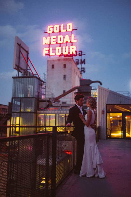 A bride and groom stand close together on an outdoor balcony at dusk, with the historic, neon-lit GOLD MEDAL FLOUR sign glowing above an industrial-style building in the background. by Justin Salem Meyer