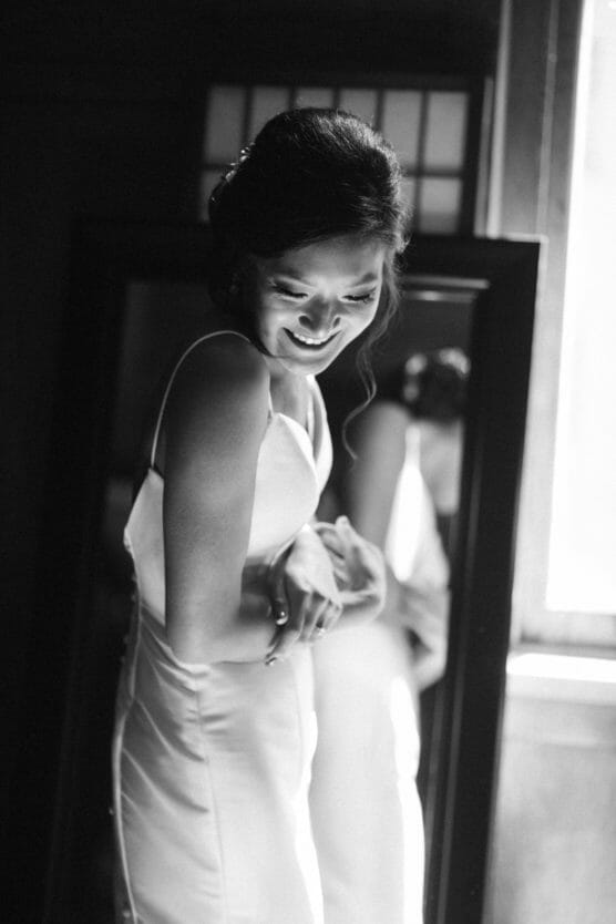 A woman in a white dress stands smiling and looking down, illuminated by natural light from a nearby window. Her reflection appears in a mirror behind her. The image is in black and white. by Justin Salem Meyer