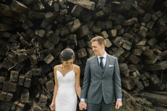 A couple dressed in wedding attire holds hands and stands in front of a large stack of weathered wooden logs. The bride looks down, smiling, while the groom gazes at her with a gentle expression. by Justin Salem Meyer
