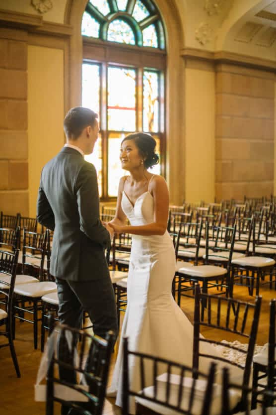 A bride in a white dress smiles and holds hands with a groom in a suit, standing among empty chairs in a sunlit room with large stained glass windows. by Justin Salem Meyer