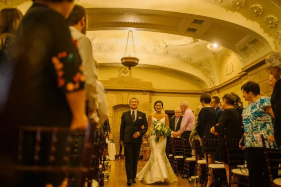 A bride in a white dress walks down the aisle with an older man in a suit, surrounded by seated guests in an elegant hall with ornate ceilings and warm lighting. by Justin Salem Meyer