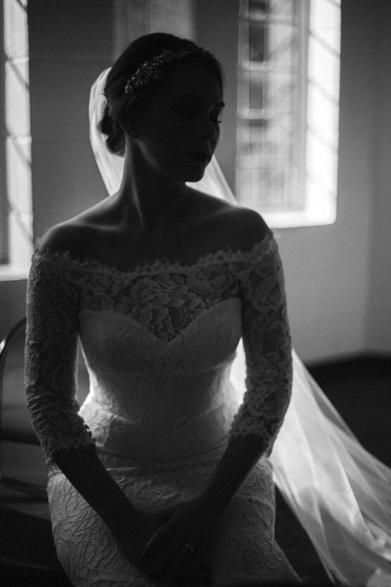 A bride in a lace off-the-shoulder wedding dress and veil sits in soft light near a window, her face and features mostly in shadow, creating a dramatic and elegant silhouette. by Justin Salem Meyer