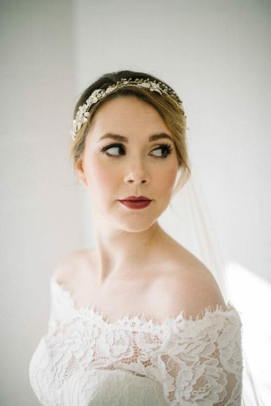 A bride in an off-the-shoulder lace wedding dress with a gold and pearl headpiece, looking to the side with soft, natural light illuminating her face. by Justin Salem Meyer