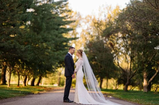 A bride and groom stand face to face on a tree-lined path, holding hands. The bride wears a long white dress and veil, while the groom is in a dark suit. Sunlight filters through the surrounding trees. by Justin Salem Meyer