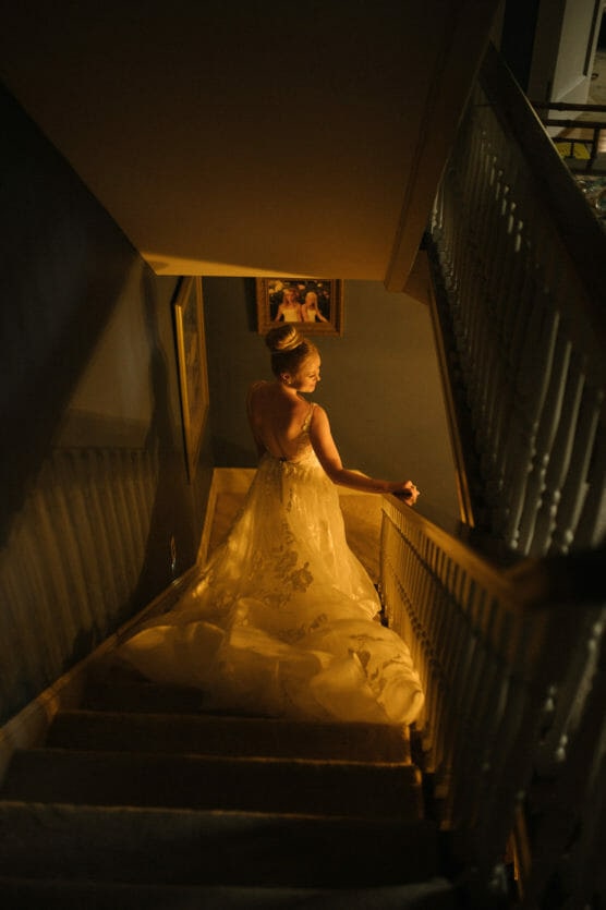 A bride in a flowing, embroidered gown stands on a dimly lit staircase, holding the railing and looking over her shoulder, with soft golden light illuminating her and framed pictures on the wall. by Justin Salem Meyer